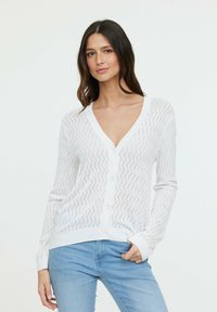 Cardigan en tricot blanc avec un motif en vagues, col en V et fermeture à boutons. Les manches sont longues, avec des poignets côtelés et une coupe décontractée.