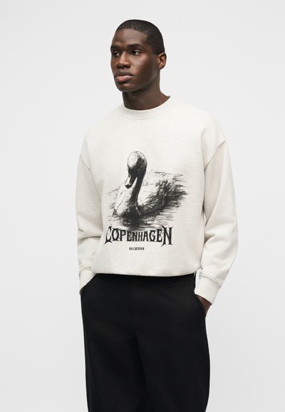 Άνδρας που φοράει μπεζ φούτερ με μαύρο γραφικό κύκνου και το κείμενο "Κοπεγχάγη", συνδυασμένο με μαύρο παντελόνι, στέκεται μπροστά σε λευκό φόντο.