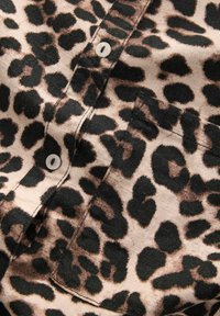 Leopardenmusterstoff mit schwarzen Flecken auf einem helleren beigen Hintergrund. Knopfdetail an einem klassischen Kragen-Design mit einer sichtbaren Tasche.