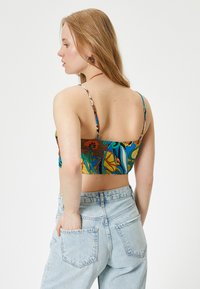 Bralette colorido con estampado y tirantes finos, que presenta un vibrante diseño floral sobre un fondo marrón y azul, combinado con unos jeans de tiro alto.