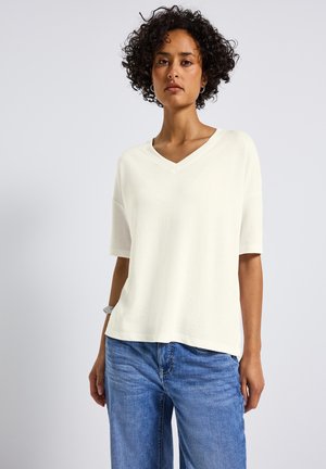 Cremfarbener V-Ausschnitt-T-Shirt mit kurzen Ärmeln, aus weichem Stoff. Einfaches Design, lockere Passform, kombiniert mit blauen Jeans.
