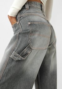Gros plan d'une personne portant un jean denim bicolore gris taille haute avec de grandes poches arrière et une main dans la poche avant, un haut blanc à manches longues visible.