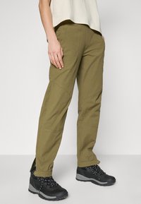 Olijfgroene buitenbroek met een rechte snit, voorzien van zichtbare stiksels en kniestukken. Gepaard met zwarte wandelschoenen met veters.