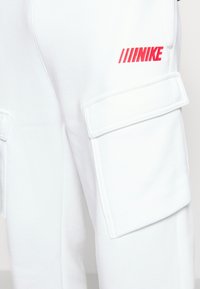 Vita cargo byxor gjorda av mjukt material, med flera fickor och en röd Nike-logotyp på sidan.