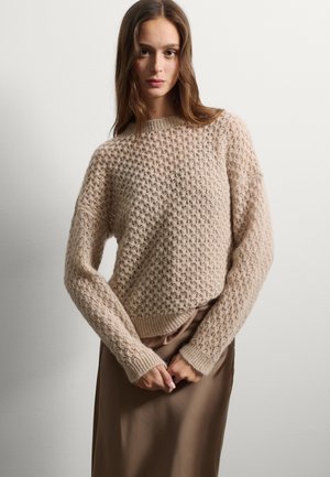 Pull en tricot beige avec un motif texturé, des poignets et un ourlet côtelés, associé à une jupe en satin marron, mettant en avant une apparence douce et confortable.