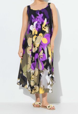 Kobieta ubrana w bezrękawnikową, luźną sukienkę do połowy łydki z dużym abstrakcyjnym kwiatowym wzorem w kolorach fioletowym, beżowym, żółtym i czarnym, do tego sandały w kwiaty.