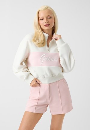 Blondynka z zamkniętymi oczami ma na sobie biało-różowy sweter zapinany na zamek z logo "Guess" i fakturowane różowe szorty na białym tle.