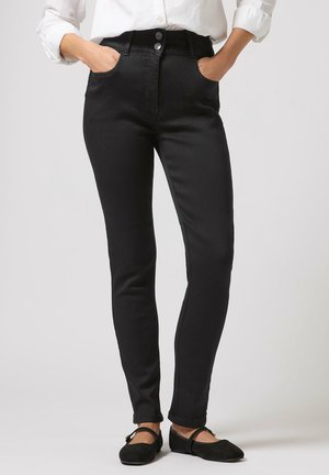 Person trägt schwarze, eng anliegende Hose mit Knopfdetail, weißes, eingestecktes Hemd und schwarze Mary Jane Ballerinas, Hände in den Taschen.