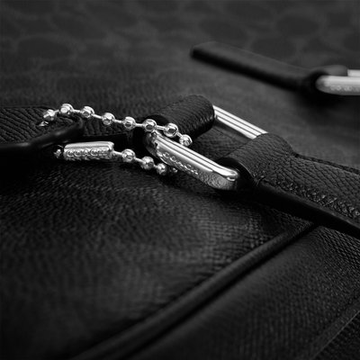 Gros plan sur cuir noir texturé avec un curseur de fermeture éclair en métal argenté et une chaîne à billes gravée du nom de la marque "Coach."