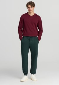 Bordowy sweatshirt z okrągłym dekoltem i długimi rękawami, zestawiony z ciemnozielonymi dresami; oba wykonane z miękkiej, teksturowanej tkaniny. Całość uzupełniają białe sneakersy.