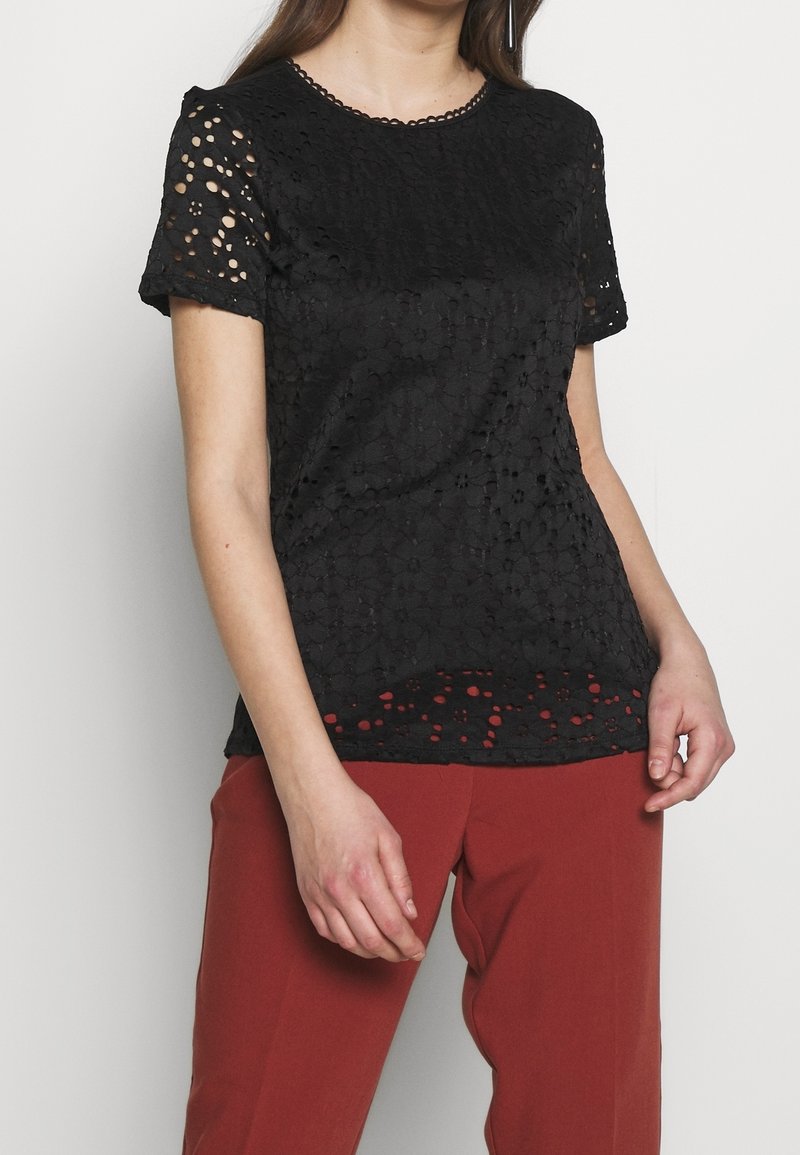 Haut en dentelle noire avec un col rond et des manches courtes, présentant un motif floral texturé et des cercles ajourés sur tout le tissu.