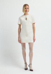 Vrouw met kort blond haar, gekleed in een crème mini-jurk met korte mouwen, met een patroon op doorschijnende panty’s en donkere puntige hakken, staande op een witte achtergrond.