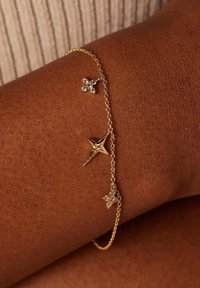 Gouden kettingarmband met stervormige en kruisvormige bedels met heldere versieringen, tegen een donkere huidskleur.