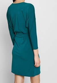 Teal omslagklänning med trekvartslånga ärmar. Mjuk, slät tyg med midjebälte och lätt utsvängd nederkant. Minimalistisk design, odekorerad.