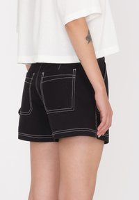 Zwarte shorts met witte stiksel, voorzien van twee achterzakken en een klein logo. De stof lijkt glad en gestructureerd.