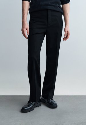 Osoba oblečená v čiernych nohaviciach s rovnými nohavicami a čiernych zaväzovacích topánkach, stojaca na sivom podlahe pred jednoduchým sivým pozadím.