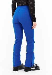 Blauwe skibroek met een strakke pasvorm, voorzien van een hoge tailleband en ritsen bij de pijpen. Gecombineerd met zwarte skibotten voor wintersport.