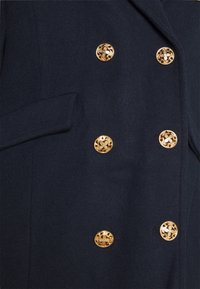 Cappotto di lana blu scuro con sei bottoni decorativi dorati a forma di fiore disposti in modo doppiopetto e una tasca a fessura orizzontale sul lato sinistro.