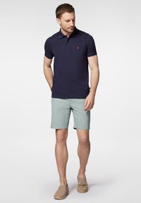 Granatowa koszulka polo z czerwonym logo, połączona z jasnymi zielonymi szortami. Buty to beżowe espadryle, materiał wydaje się gładki i swobodny.