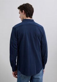 Camisa azul marino de manga larga con cuello con solapa, tejido texturizado, dobladillo curvado y puños con botones, usada sobre unos vaqueros azul claro.