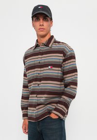 Bruin en beige gestreepte langemouwen blouse met blauwe accenten en een borstzak; heeft een logo patch op de zak. Grijze pet met logo.