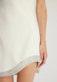 Robe blanche avec un ourlet arrondi, ornée d'une bordure en bijoux argentés sur le bas. Tissu lisse, design ajusté et longueur courte.