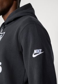 Čierna mikina s kapucňou a vačkou v štýle klokania, s textúrovaným povrchom. Obsahuje biele logo Nike na rukáve. Kapucňový dizajn, voľný strih.
