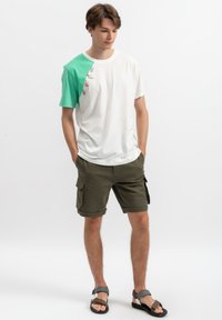 Witte t-shirt met een groene linkermouw, voorzien van een kleurrafische afbeelding. Gecombineerd met olijfgroene cargoshorts en bruine sandalen.