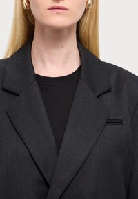 Czarna, dopasowana marynarka z ostrymi klapami, teksturowanym materiałem i niewielkim detalem kieszonki. Noszona na czarnym topie z okrągłym dekoltem. Minimalistyczny design.