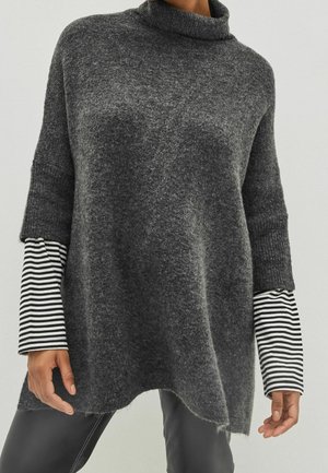 Personne portant un pull à col roulé gris ample par-dessus une chemise à manches longues rayée noir et blanc et un pantalon foncé, debout devant un fond uni.