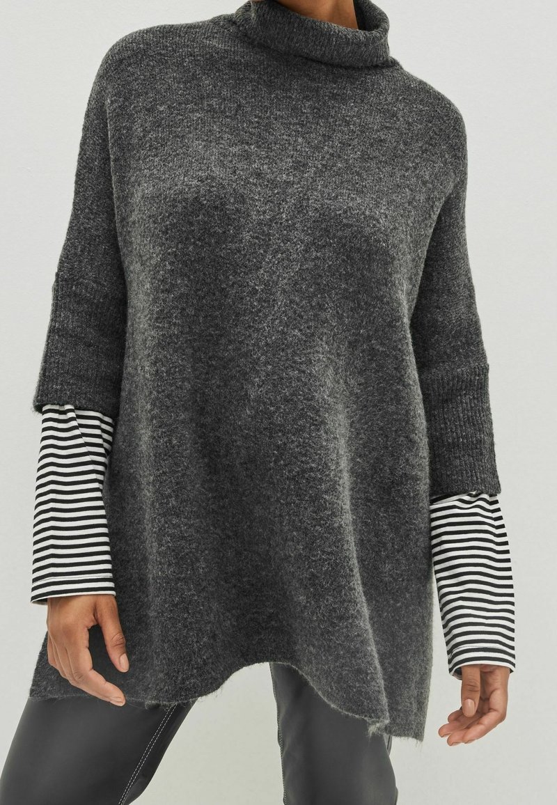 Personne portant un pull à col roulé gris ample par-dessus une chemise à manches longues rayée noir et blanc et un pantalon foncé, debout devant un fond uni.