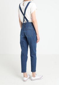 Jeansbyxor med korsettselar, mörkblå färg, smalnade ben, två bakfickor, kombinerade med en vit t-shirt och vita sneakers.