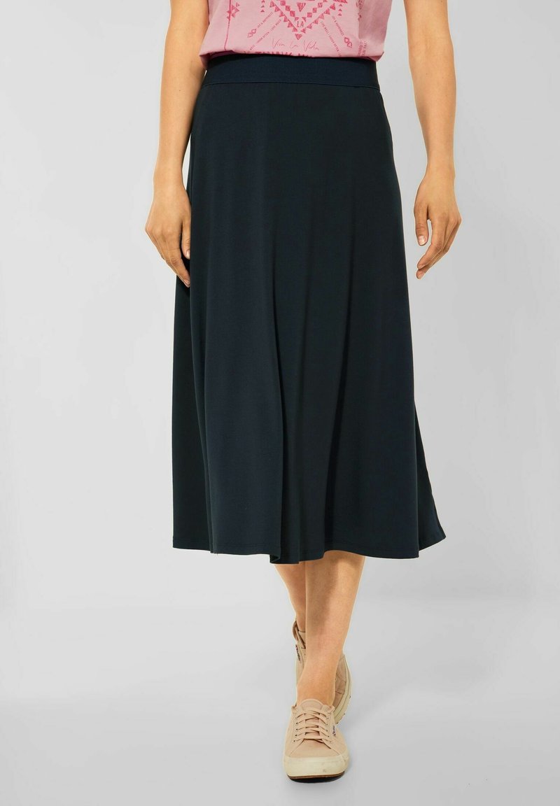 Femme portant une jupe midi noire, un haut rose à motifs et des baskets beiges, debout sur un fond uni gris clair.