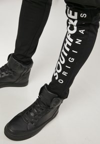 Svart høye joggesko med teksturert lær, svarte bukser med hvit "SOUTHPOLE ORIGINALS" tekst langs siden, slim fit design.