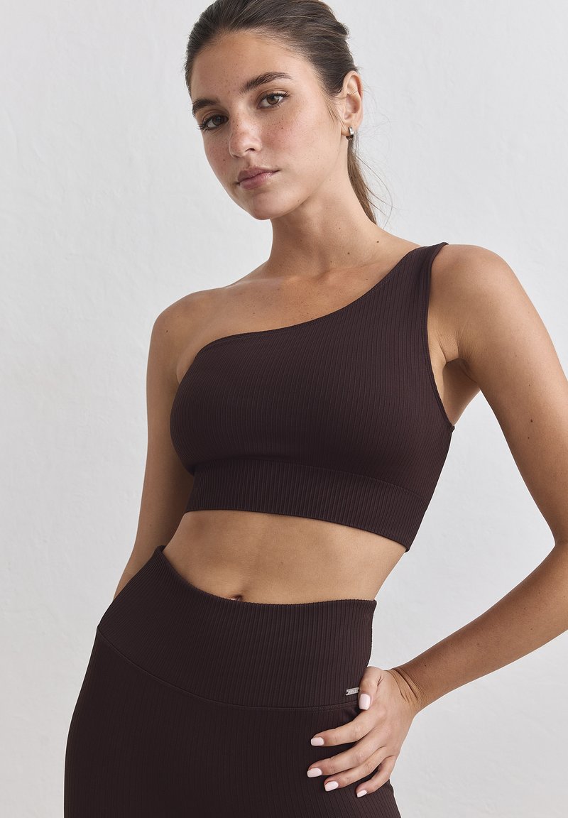 Γυναίκα που φοράει σκούρο καφέ ριμπ πλεκτό crop top με έναν ώμο και ταιριαστά κολάν με ψηλή μέση, ποζάρει με το χέρι στη μέση μπροστά σε απλό φόντο.