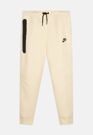 Pantalons de survêtement crème clair avec un cordon de serrage noir ajustable, une poche zippée sur le côté et un logo Nike noir sur le côté gauche. Texture de tissu lisse.