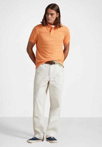 Πορτοκαλί πουκάμισο πόλο με την δύο κουμπιών, συνδυασμένο με ανοιχτό μπεζ παντελόνι και μπλε αθλητικά παπούτσια. Χαλαρή εμφάνιση με ελάχιστες σχεδιαστικές λεπτομέρειες.
