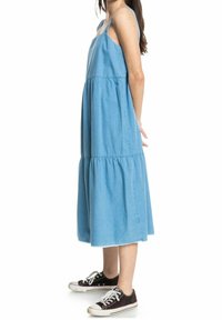 Blauwe denim midi-jurk met spaghettibandjes, gelaagd ontwerp en een losse pasvorm, met ruwe zoomranden en gecombineerd met zwarte sneakers.