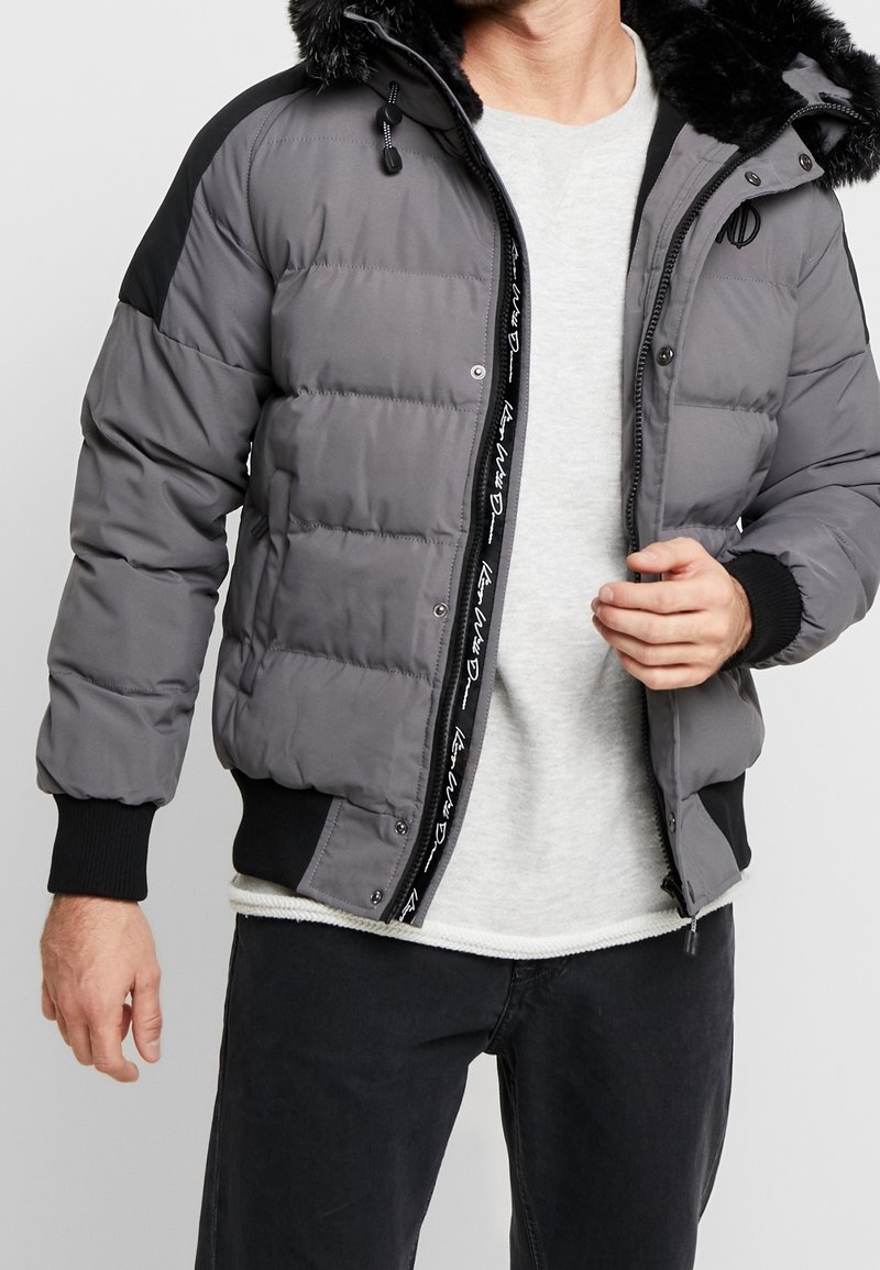 Fyllig grå jacka med svart huva och mudd. Har en vävd inneretikett och en dragkedjestängning med tryckknappar för säker stängning.