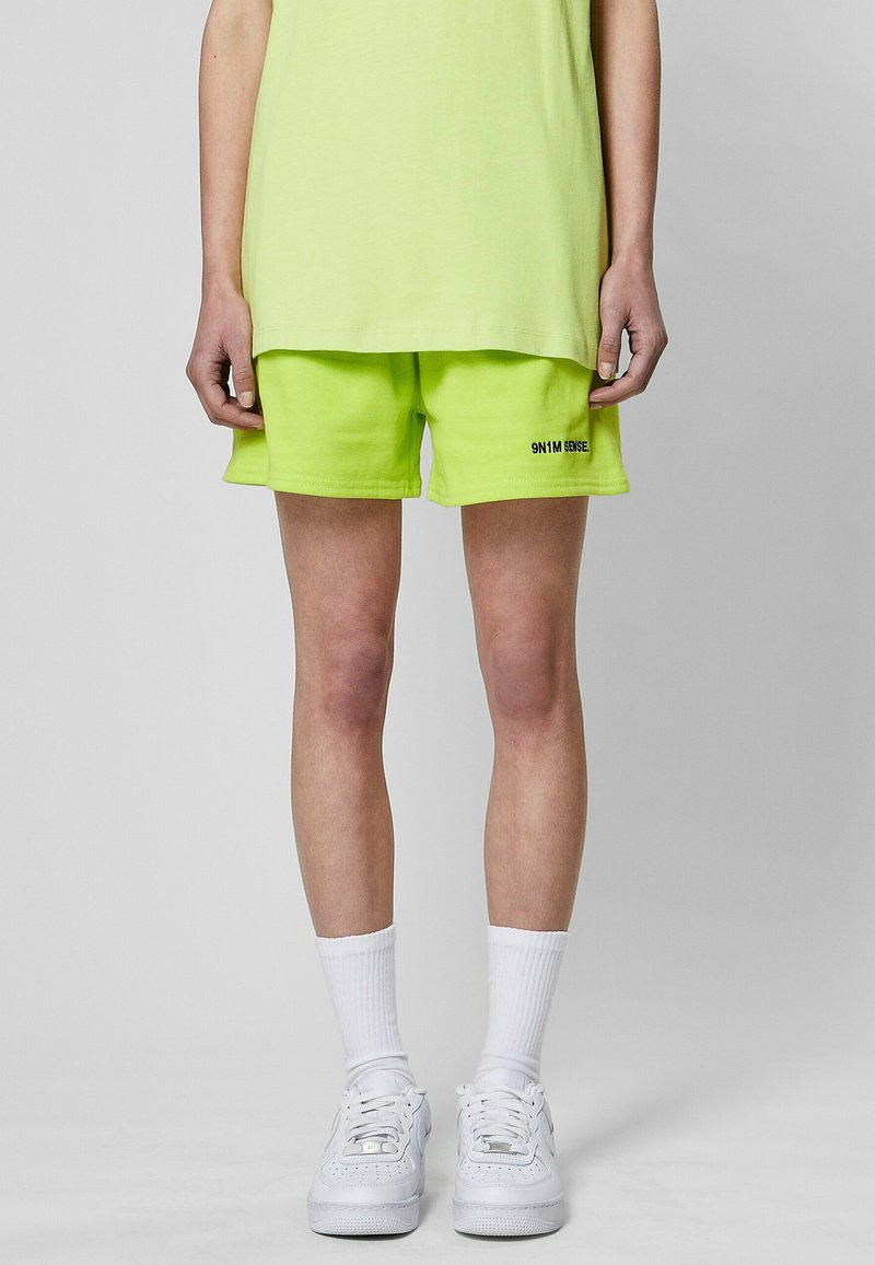 Limegrüne Baumwollshorts mit lässigem Schnitt, mit einem schwarzen aufgedruckten Logo am linken Bein, getragen mit weißen Turnschuhen und gerippten Socken.