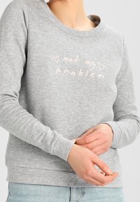 Grå sweatshirt med långa ärmar och rundad halsringning. Har ljusrosa broderad text som säger "inte mitt problem" med hjärtaccenter.