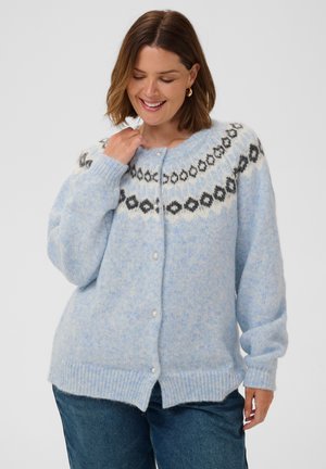 Femme portant un cardigan tricoté bleu clair avec un yoke à motif noir et blanc, des boutons en perle et un jean bleu, souriante et regardant vers le bas.