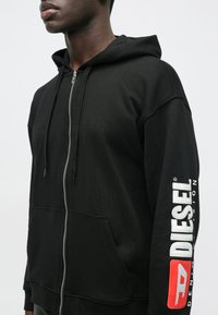 Muškarac nosi crni duksericu s patentnim zatvaračem i prednjim džepovima, a na lijevoj rukavu se nalazi "DIESEL" logo u bijeloj i crvenoj boji, u pozadini bijela.