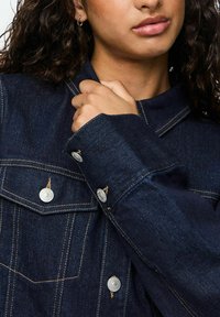Gros plan sur une personne portant une veste en denim bleu foncé avec des boutons-pression argentés, tenant le col de la veste près de son cou.