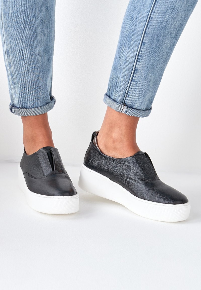 Black leather slip-on shoes with textured finish, featuring a white platform sole and elastic side panels. Worn with cuffed light denim jeans.