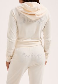 Set di tuta color crema composto da giacca con cappuccio e dettagli in strass sulla schiena, abbinata a pantaloni con ricamo in strass "Juicy".
