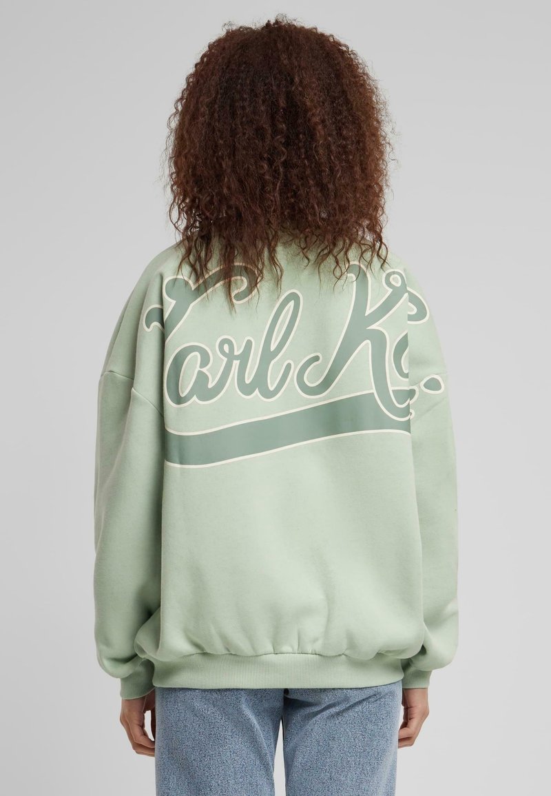 Personne aux cheveux bouclés portant un sweat-shirt ample vert clair avec un grand texte en écriture blanche au dos, assorti à un jean bleu.