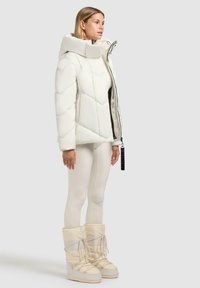 Witte pufferjack met een hoge kraag en gewatteerd patroon, gecombineerd met off-white legging en snowboots tot de enkels met veters.