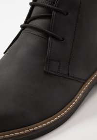 Bottine en cuir noir avec une texture lisse, bout rond et coutures contrastantes. Dispose d'un système de laçage et d'une semelle robuste.