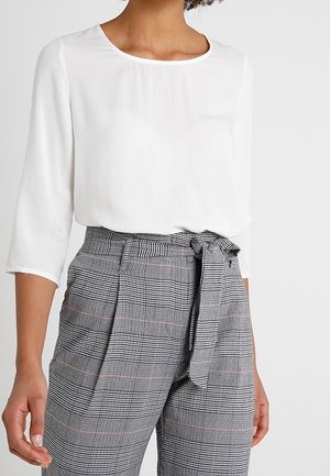 Frau trägt eine weiße Dreiviertelarmbluse, die in hoch taillierte graue karierte Hose mit gebundenem Gürtel in der Taille gesteckt ist.